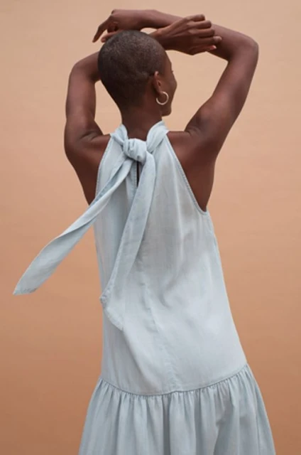 Back view of an adult woman with short hair wearing a light blue dress tied at the neck against a neutral brown background.