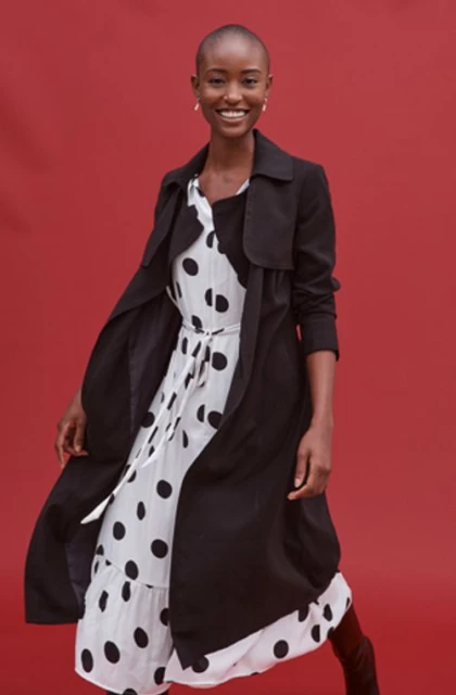 Smiling adult Black female in black trench coat over polka dot dress against red studio backdrop in medium full shot.