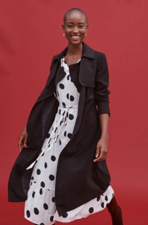 Smiling black model with short hair wearing a black trench coat over a white polka dot dress against a red background.