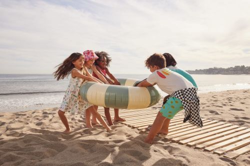 Children play on a sandy beach with a green and beige inflatable tube, with the ocean and sky in the background on a sunny day.