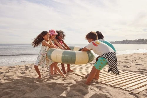 Children play on a sandy beach with a green and beige inflatable tube, with the ocean and sky in the background on a sunny day.