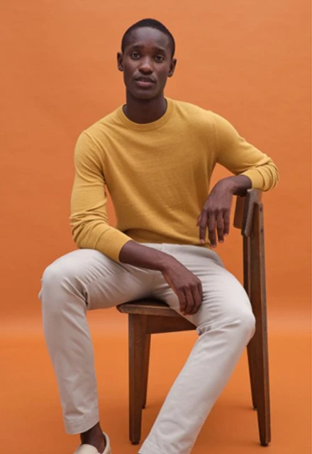 Portrait of a Black man in a mustard sweater and beige pants sitting on a wooden chair against an orange background in a studio setting.