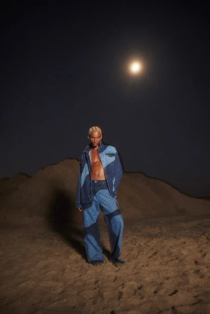 A blond male fashion model in a denim outfit poses in a desert environment at night under the moonlight in a full-length shot.