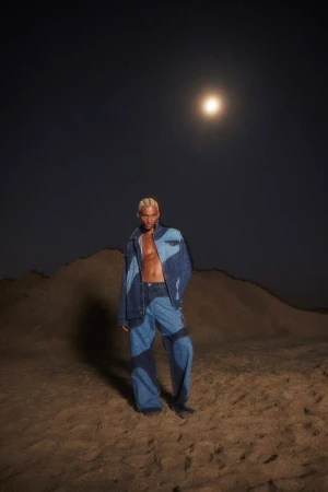 A blond male fashion model in a denim outfit poses in a desert environment at night under the moonlight in a full-length shot.