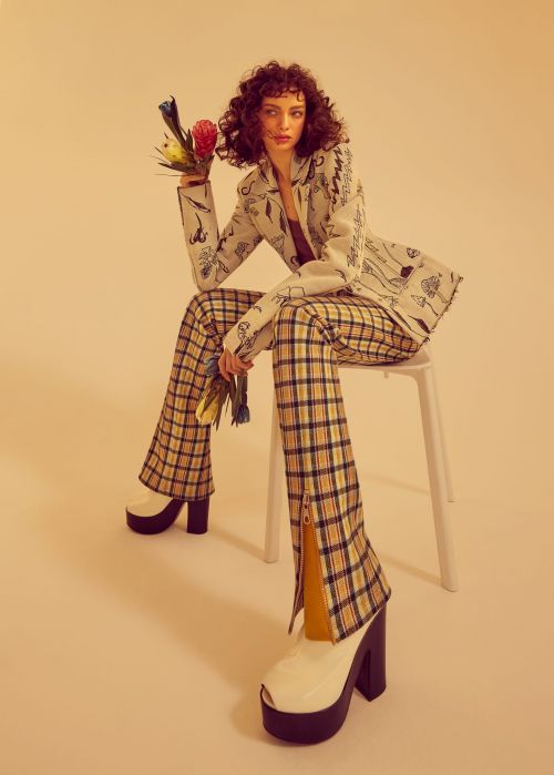 Fashion shot of a brunette woman in a patterned suit and platform shoes, sitting on a stool and holding flowers in a studio.