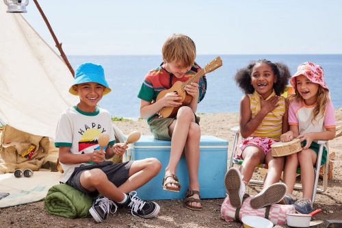 Four smiling children enjoy a beach picnic, one playing ukulele, one with maracas, another playing a toy drum near a tent on a sunny day.
