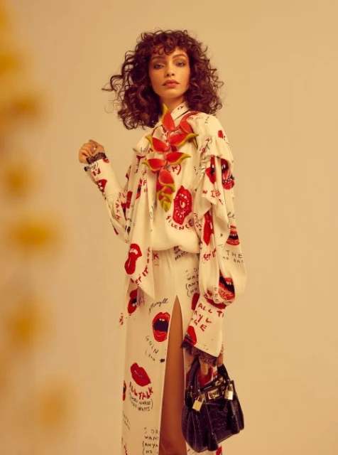 Woman in a printed white dress and watermelon necklace poses with a black purse against a neutral background in a fashion shot.