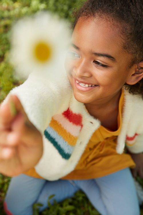 Young girl smiles, holding a daisy in a grassy outdoor setting, wearing a colorful striped cardigan and yellow top.