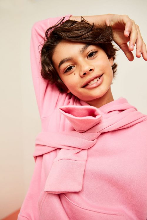 A young teen smiles in a cheerful indoor portrait, wearing a pink hooded sweatshirt tied around the neck in front of a neutral backdrop.