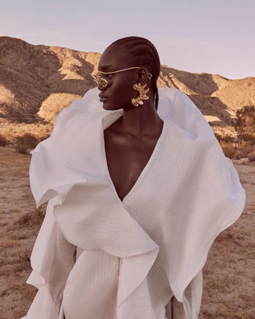 Woman in a ruffled white dress and glasses poses in a desert landscape with soft lighting, showcasing fashion and beauty.