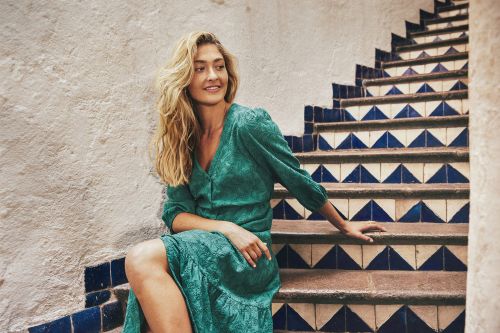Adult female in a patterned green dress sitting on outdoor stairs with a textured wall in the background, looking off into the distance.