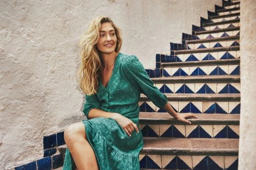 Adult female in a patterned green dress sitting on outdoor stairs with a textured wall in the background, looking off into the distance.
