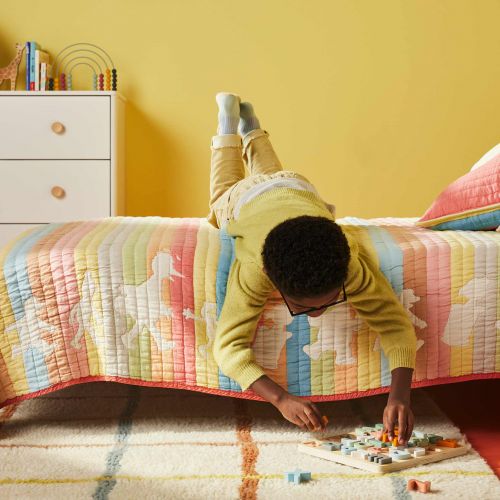 Young child lies upside down on bed, wearing glasses, playing with a board game on a striped rug in a cheerful, yellow bedroom.
