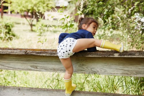 A young child wearing patterned diaper and yellow boots climbs on a rustic wooden fence in a grassy outdoor setting with trees.