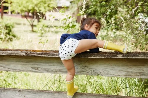 A young child wearing patterned diaper and yellow boots climbs on a rustic wooden fence in a grassy outdoor setting with trees.