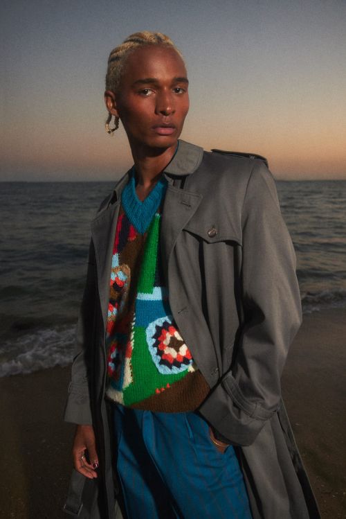 A stylish dark-skinned male model with blonde braids poses on a beach at dusk, wearing a colorful vest, trench coat, and blue pants.