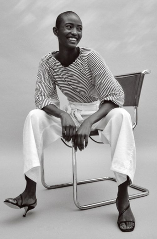 Black woman with a shaved head wearing a striped top and white trousers seated on a chrome chair in Los Angeles