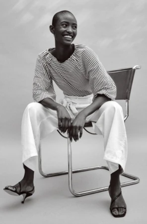 Black woman with a shaved head wearing a striped top and white trousers seated on a chrome chair in Los Angeles