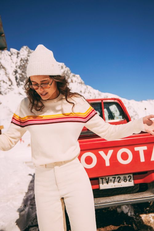 Woman in winter fashion stands by a red pickup truck in a snowy mountain setting, wearing a white beanie, sweater, and sunglasses.