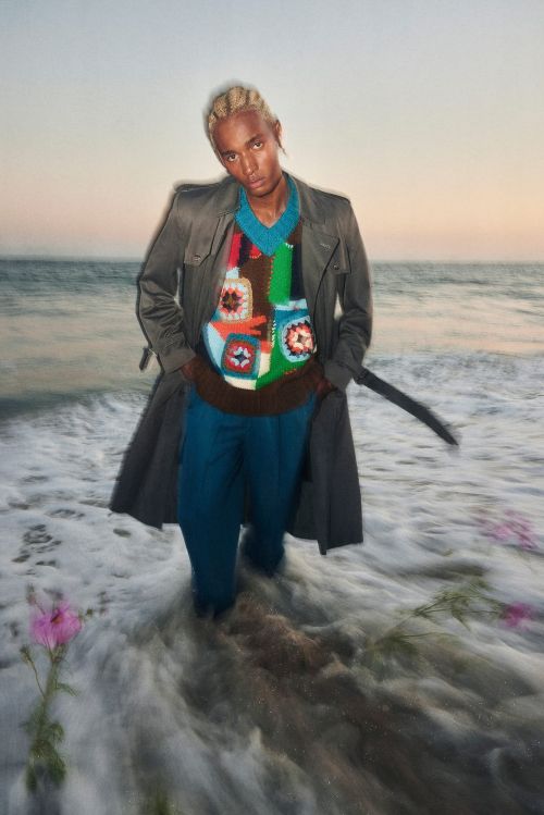 Man in patterned sweater and trench coat standing in ocean waves, looking at camera. Water and sunset sky background.
