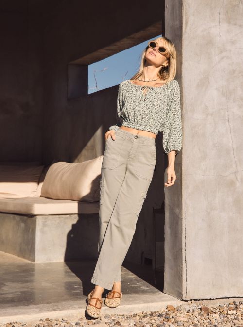 A fashionable woman in sunglasses poses in casual clothing near a concrete structure, bathed in sunlight. She wears a floral top and cargo pants.