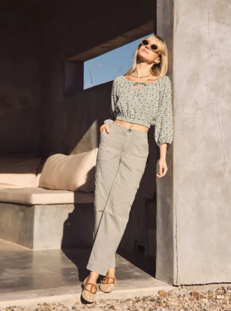 A fashionable woman in sunglasses poses in casual clothing near a concrete structure, bathed in sunlight. She wears a floral top and cargo pants.