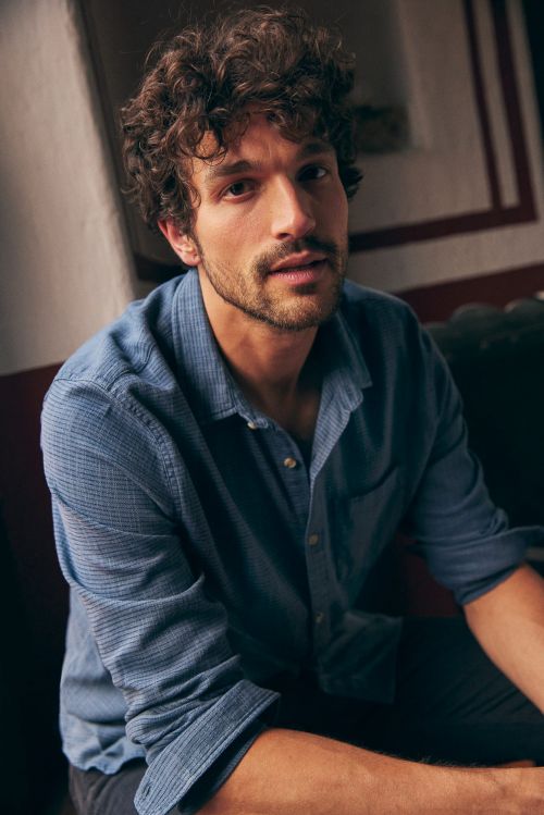 Portrait of a man with curly brown hair and a blue button-down shirt, sitting indoors with soft, natural light.