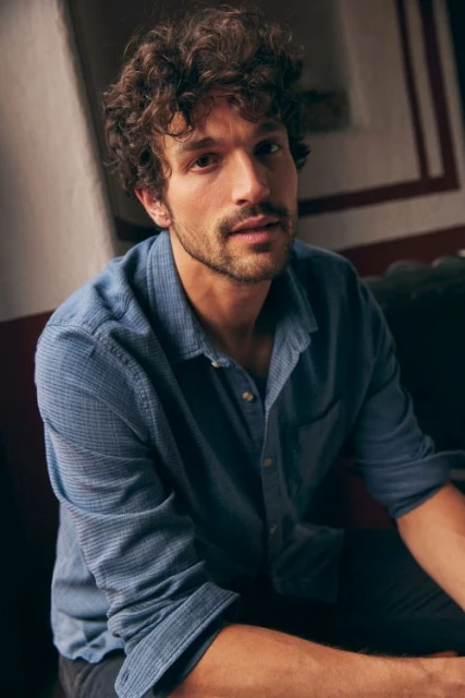 Portrait of a man with curly brown hair and a blue button-down shirt, sitting indoors with soft, natural light.
