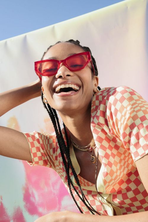 A happy Black woman wearing sunglasses and a checkered shirt smiles in front of a backdrop with pink and blue highlights, outdoors.