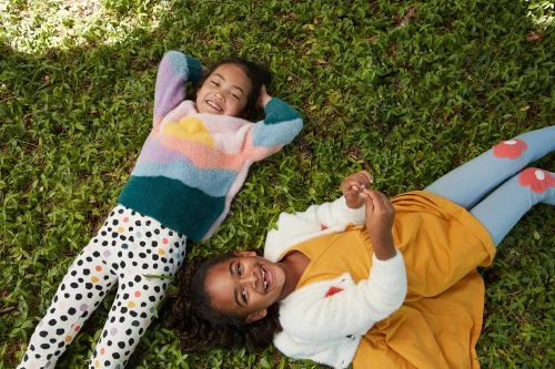 Two smiling girls lie on green grass in sunlight wearing colorful sweaters and leggings with polka dot and mushroom patterns. 