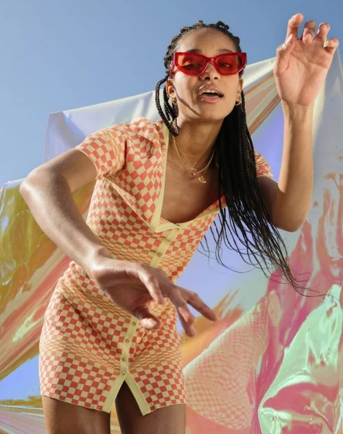 Woman with braided hair wears orange checkerboard mini dress and red sunglasses against blue sky and iridescent sheet backdrop.