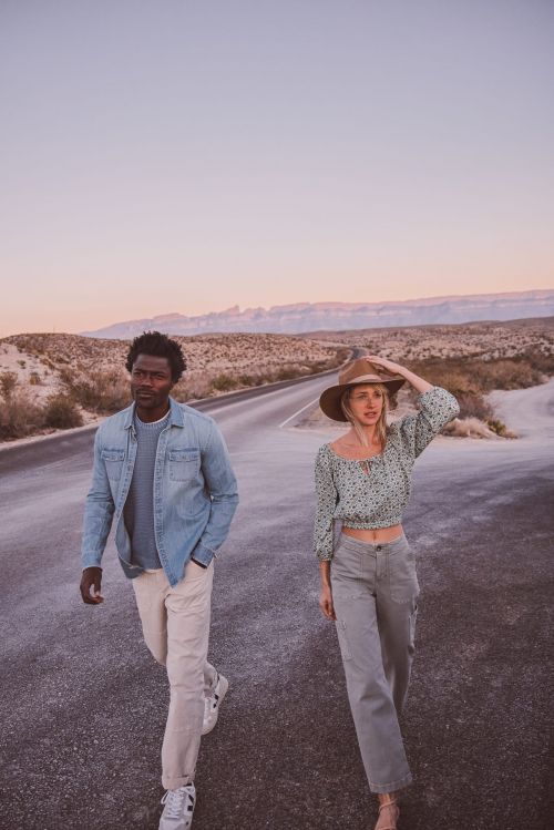 A man and woman walk on a desert road during golden hour. The woman wears a hat and green blouse, while the man has a denim shirt.