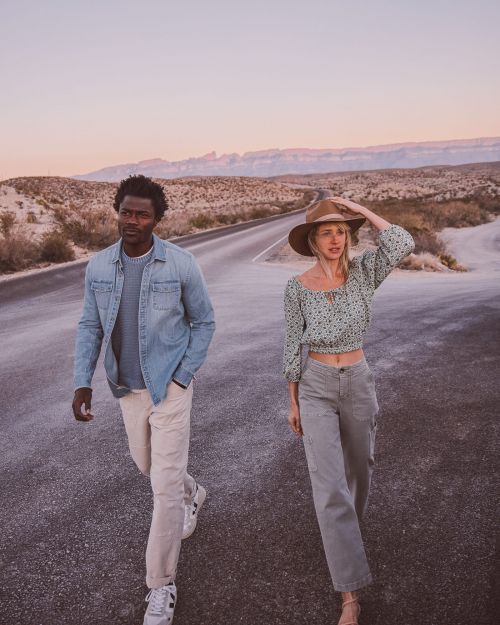 A couple walks on a desert road at sunset. A Black man and a White woman model casual fashion in a scenic outdoor landscape.