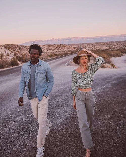 A couple walks on a desert road at sunset. A Black man and a White woman model casual fashion in a scenic outdoor landscape.