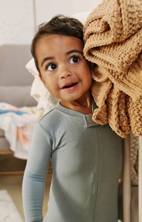 A young child in a blue jumpsuit stands next to a pile of blankets, looking up and to the side in an indoor setting with natural lighting.