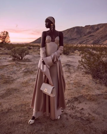 Fashion portrait of a Black woman in a flowing gown, standing in a desert landscape at dusk, accessorized with a handbag and jewelry.