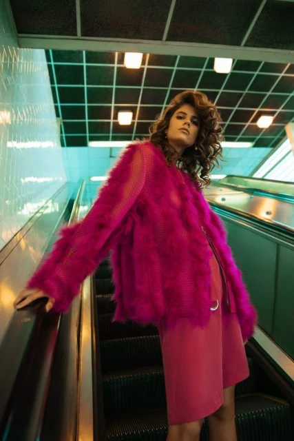 Indoor shot of a woman in a pink coat and dress posing on an escalator with a patterned ceiling and turquoise wall in the background.