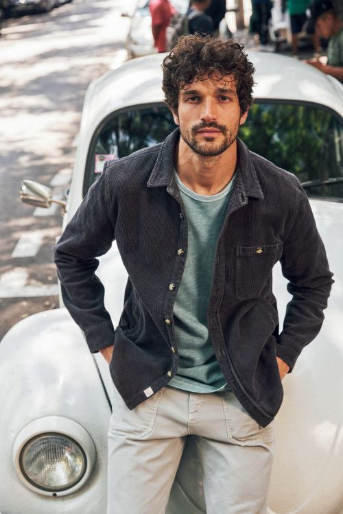A man with dark hair and a light blue shirt and dark jacket stands next to a white car on a city street, with a blurred background.