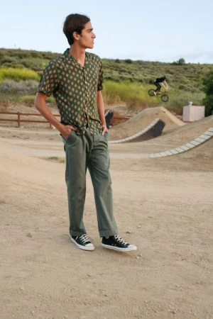 Man in patterned shirt stands on dirt ground as a second man jumps a bike on a distant ramp with natural grassy landscape backdrop.
