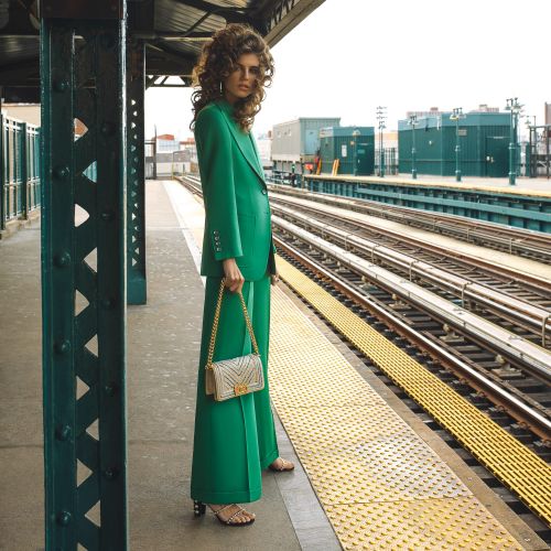 Woman in a green suit with a handbag stands on an outdoor train platform with yellow safety tiles. Urban fashion editorial shot.