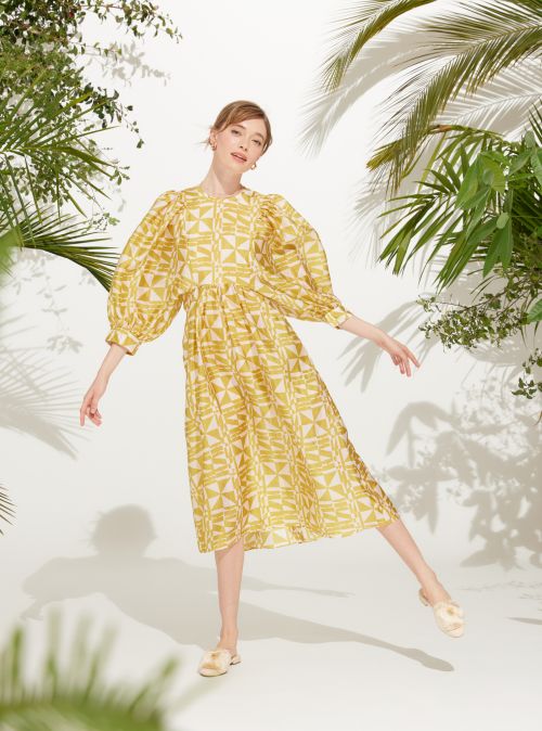 A fashionable adult woman wearing a patterned yellow dress with puffy sleeves poses in a studio with tropical foliage and palm leaf shadows.