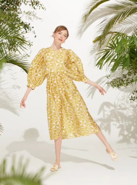 A fashionable adult woman wearing a patterned yellow dress with puffy sleeves poses in a studio with tropical foliage and palm leaf shadows.