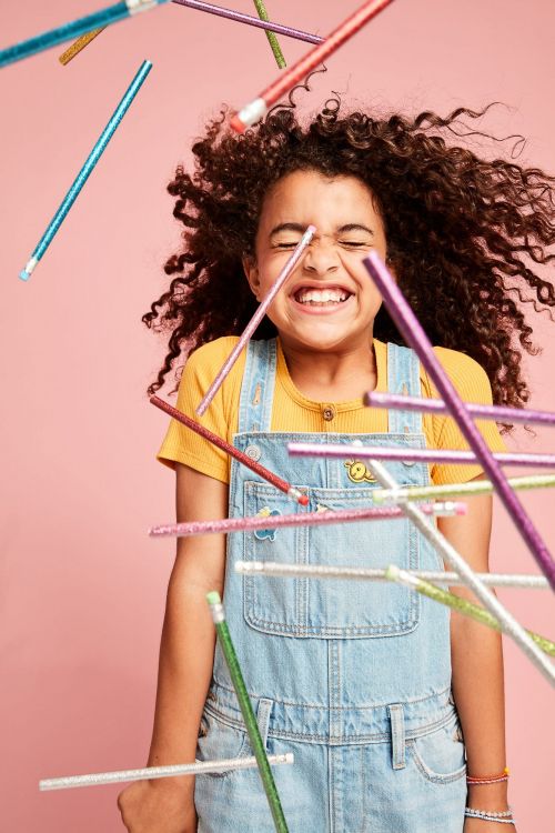 A young girl smiles widely surrounded by floating pencils in a pink studio setting. The image captures a playful and joyful moment.