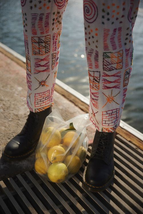 Person stands on a dock in black boots and patterned pants, holding a plastic bag full of yellow lemons in bright outdoor light.