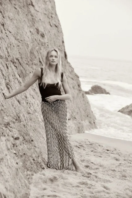 Monochrome portrait of a blond woman in a long patterned skirt posing next to a rock formation at the beach with ocean in background.