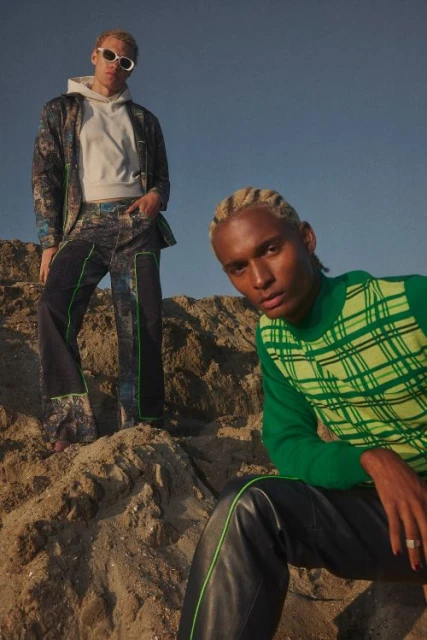 Two fair-skinned men pose on a sand dune. One wears sunglasses, a suit, and a white hooded sweatshirt; the other, a green patterned sweater.
