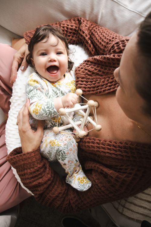 Adorable baby girl in floral-print outfit smiles while lying in someone's arms, playing with a wooden geometric toy.