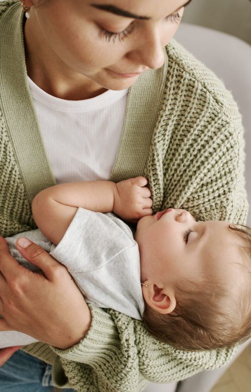 A woman holds a sleeping baby in her arms, both are wearing casual clothing and the soft lighting adds a gentle touch to the scene.