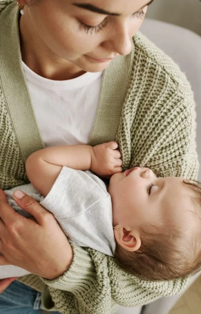A woman holds a sleeping baby in her arms, both are wearing casual clothing and the soft lighting adds a gentle touch to the scene.