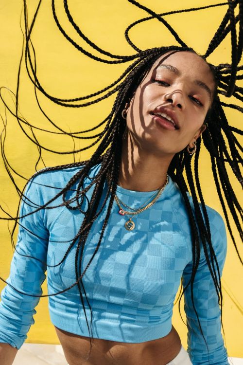 A young woman with braids poses in front of a yellow background, wearing a blue long-sleeved crop top and gold necklaces in a studio shot.